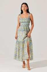 A woman is wearing a sleeveless, ankle-length dress with spaghetti straps tied at the shoulders. The dress features a fitted, smocked bodice and a flowy skirt with a slit up one side. The fabric has a yellow, blue, and green floral pattern. She is accessorized with earrings, a bracelet, and rings, and is wearing open-toe heels.